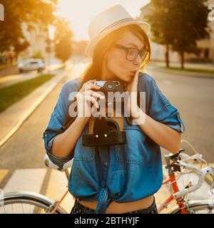 femme élégante en chapeau près du vélo sous le soleil du matin avec appareil photo extérieur Banque D'Images