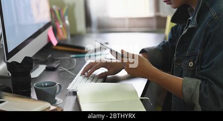 Image rognée d'un homme de designer tenant/regardant un smartphone à la main tout en tapant sur le clavier et assis devant un écran blanc d'ordinateur moniteur Banque D'Images