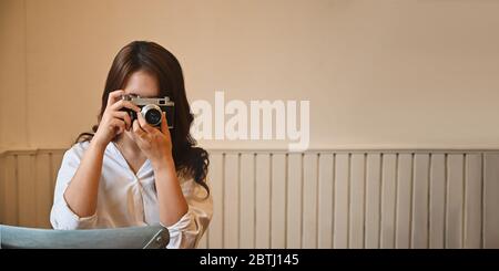 Belle femme photographiant vers l'appareil photo avec un appareil photo vintage tout en étant assise dans un salon confortable. Banque D'Images