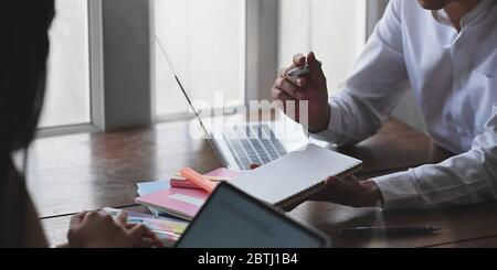 Image rognée d'un homme d'affaires prenant des notes en étant assis devant un ordinateur portable et de son collègue au bureau en bois sur le salon gagne Banque D'Images