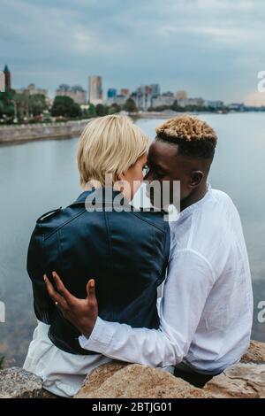 Le couple interracial est assis sur des rochers et se hople contre le fond de la rivière et de la ville. Concept de relations d'amour et d'unité entre les différentes races humaines. Banque D'Images