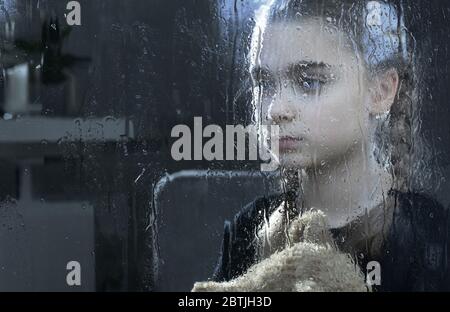 Dépression chez les enfants. Caucasienne fille avec ours en peluche derrière la fenêtre humide de pluie. Photo conceptuelle. Banque D'Images