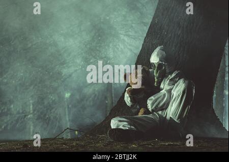 Hommes en blanc Hazmat Suite et masque à gaz se posant sous l'arbre avec sa fille Teddy Bear Toy à côté de lui. Nuit sombre dans la forêt couverte de fumée. Banque D'Images