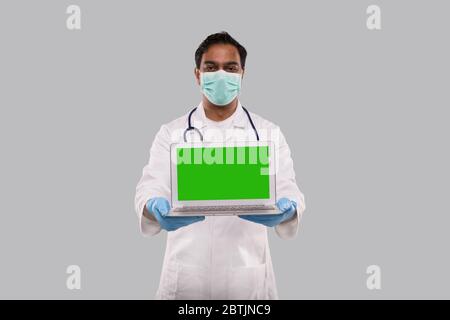 Médecin montrant un ordinateur portable écran vert isolé portant un masque médical et des gants. Indian Man Doctor avec un ordinateur portable en mains. Concept de médecine en ligne Banque D'Images