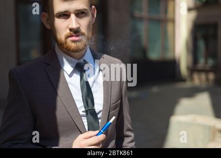 iqos de cigarettes électroniques. Portrait d'un jeune homme barbu de vingt-cinq ans, debout par le mur texturé du bâtiment, fume une cigarette. Banque D'Images