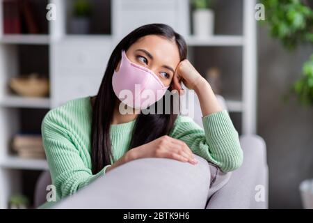 Photo de joli mais triste virus corona malade patient homey asiatique femme assis canapé regarder fenêtre rêveuse manquant aller à l'extérieur besoin de garder l'auto-isolement Banque D'Images