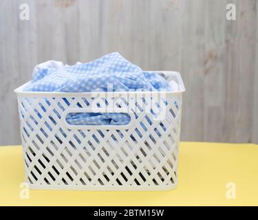 Chemises blanches bleues vierges dans un panier en plastique blanc sur une table jaune sur fond gris. Blanchisserie, nettoyage à la maison, propreté et fraîcheur Banque D'Images