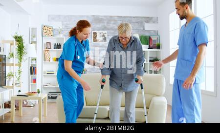 Femme âgée avec des béquilles qui obtient de l'aide pour que l'infirmière et le médecin de sexe féminin se tiennent debout sur les jambes du canapé Banque D'Images