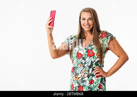 Jeune femme souriante dans une robe colorée regardant volontiers dans l'appareil photo prendre des photos sur un téléphone portable sur fond blanc isolé Banque D'Images