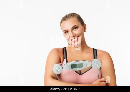 Portrait d'une femme souriante avec un poids excessif tenant des balances dans les mains tout en regardant avec plaisir dans l'appareil photo sur fond blanc Banque D'Images