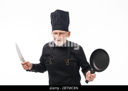 Homme vieux et attrayant avec une barbe grise dans un tablier noir et un capuchon tenant une casserole et un couteau dans les mains regardant avec soin dans l'appareil photo sur fond blanc isolé Banque D'Images