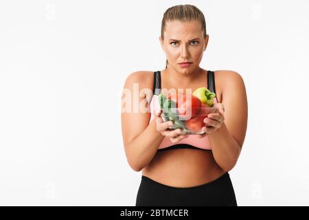 Jeune femme avec un poids excessif dans le haut sportif regardant malheureusement dans l'appareil photo tout en tenant un bol avec des légumes dans les mains sur fond blanc Banque D'Images