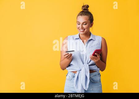 Jeune femme joyeuse en chemise joyeuse tenant la carte de crédit et le téléphone cellulaire dans les mains sur fond jaune isolé Banque D'Images