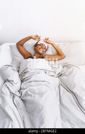 Une fille afro-américaine souriante dans un masque de sommeil s'est réveillée dans la chambre Banque D'Images