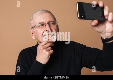 Vieil homme attrayant avec cheveux gris et barbe en lunettes et chandail prendre des photos sur téléphone portable sur fond beige isolé Banque D'Images