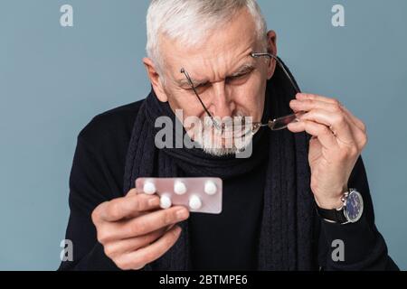 Homme malade pensif vieux avec cheveux gris et barbe dans les lunettes et foulard regardant soigneusement sur les pilules sur fond bleu isolé Banque D'Images