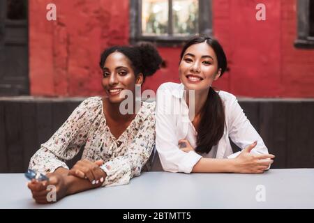 Deux belles filles souriantes se réjouieusement de regarder dans l'appareil photo tout en passant du temps ensemble dans la cour confortable du café