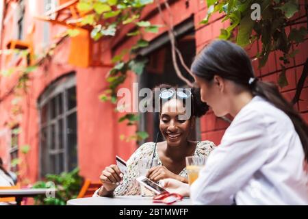 Jolies filles souriantes utilisant le téléphone portable et la carte de crédit tout en passant du temps ensemble dans la cour confortable du café Banque D'Images