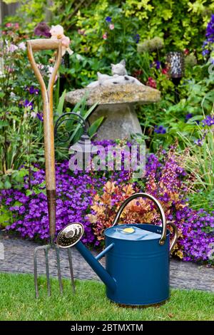 Arrosoir traditionnel en métal et fourche de jardin avec un beau jardin de style cottage en arrière-plan Banque D'Images