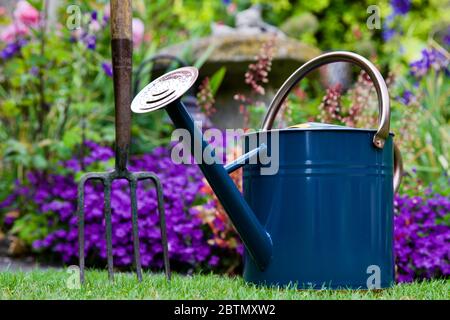 Arrosoir traditionnel en métal et fourche de jardin avec un beau jardin de style cottage en arrière-plan Banque D'Images