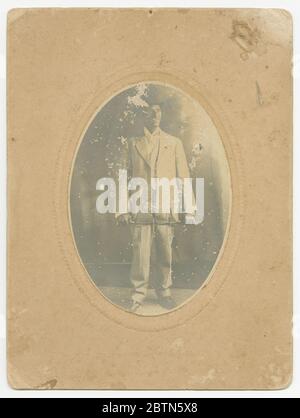 Photographie d'un homme. Cette photo en noir et blanc représente un portrait complet d'un homme. Porter un chapeau foncé, un costume de couleur claire et porter une canne. Il se tient face à la caméra, avec son regard dirigé vers la droite du spectateur. Il porte sa canne devant lui, tenant chaque extrémité de chaque main. Banque D'Images