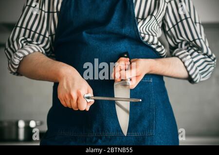 Le cuisinier, habillé d'un tablier bleu et d'une chemise rayée, est debout dans la cuisine aiguisant une lame de couteau avec une pierre à aiguiser. Banque D'Images