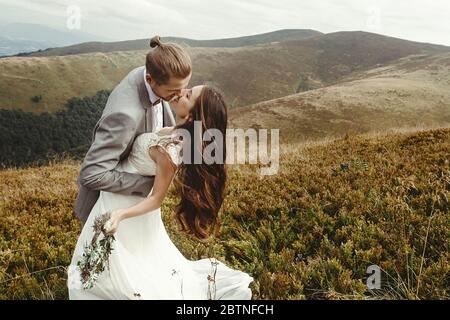 marié élégant embrassant la mariée magnifique dans la lumière du soleil, moment parfait, couple de mariage de boho, cérémonie de luxe à la montagne avec vue incroyable Banque D'Images