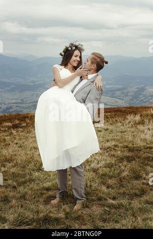 tenue élégante de marié transportant une mariée heureuse dans la lumière du soleil et souriant, moment romantique, couple de mariage de boho, cérémonie de luxe à la montagne avec v incroyable Banque D'Images