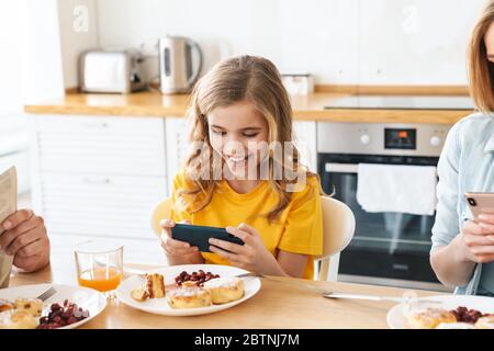 Photo de drôle jolie fille jouant au jeu vidéo tout en ayant le petit déjeuner avec sa famille dans la cuisine moderne Banque D'Images