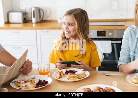 Photo de drôle jolie fille jouant au jeu vidéo tout en ayant le petit déjeuner avec sa famille dans la cuisine moderne Banque D'Images