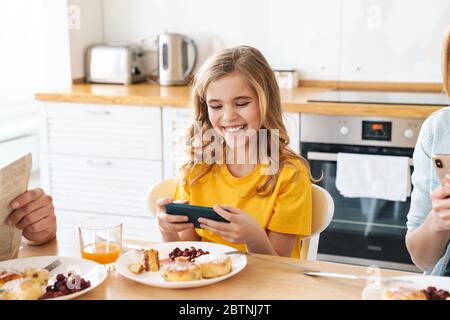 Photo de drôle jolie fille jouant au jeu vidéo tout en ayant le petit déjeuner avec sa famille dans la cuisine moderne Banque D'Images