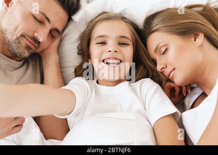 Photo de joyeuse fille caucasienne prenant photo selfie tout en étant couché avec des parents dormant dans blanc mauvais Banque D'Images