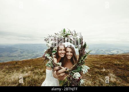 élégante demoiselle d'honneur et superbe mariée riant et regardant à travers couronne, mariage de boho, cérémonie de luxe à la montagne Banque D'Images