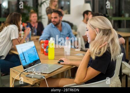 Une femme travaille sur un ordinateur portable pendant que ses collègues se rencontrent et socialisent en arrière-plan. Banque D'Images