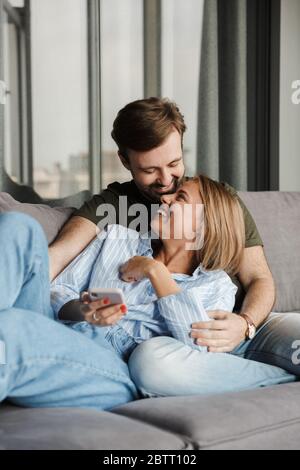 Photo de drôle beau couple gentil rire et en utilisant le téléphone mobile tout en étant assis sur le canapé à la maison Banque D'Images
