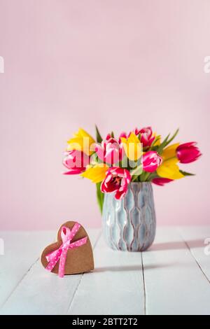 Boîte cadeau en forme de coeur avec noeud en ruban satiné et bouquet flou de fleurs de tulipe rose et jaune dans un vase sur la table en bois blanc a Banque D'Images