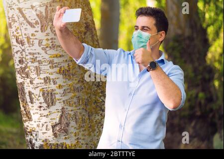 COVID-19 homme beau optimiste portant un masque chirurgical sur le visage pendant la maladie pandémique du coronavirus montrant les pouces dans le parc de la ville en faisant un appel vidéo . Banque D'Images