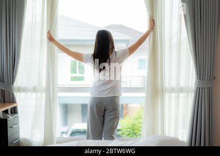 Vue arrière de l'Asian Woman se réveillant dans son lit complètement reposé et rideaux de la fenêtre d'ouverture en regardant par la fenêtre dans la chambre à la maison. Banque D'Images