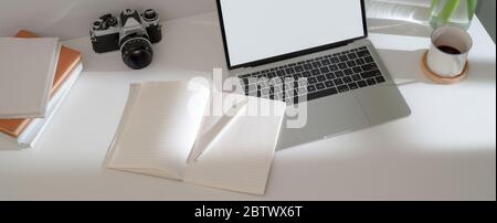 Photo courte d'un bureau simple avec ordinateur portable maquette, carnets de notes, tasse à café et appareil photo sur table blanche Banque D'Images