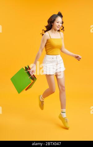 Belle jeune femme shopaholique sautant dans l'air avec des sacs de shopping sur fond orange. Banque D'Images