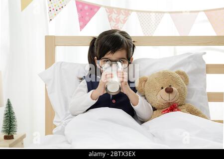 Petite fille asiatique boit du lait sur le lit en bois de sa chambre, fille Preschooler boit du lait avec du verre le jour ensoleillé, concep éducatif Banque D'Images