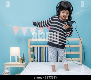 Une petite fille mignonne chantant une chanson avec un smartphone dans sa chambre, une petite fille asiatique heureuse qui écoute la musique avec un casque sur le lit, technologie c Banque D'Images