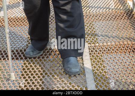 Homme tenant un bâton de marche et portant un pantalon noir et de vieilles chaussures rayées faisant un pas dur sur une surface métallique Banque D'Images