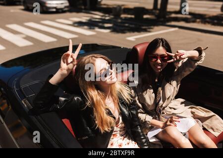 Deux belles filles en lunettes de soleil se réjouissent de passer du temps ensemble tout en conduisant une voiture cabriolet dans les rues de la ville en plein air Banque D'Images