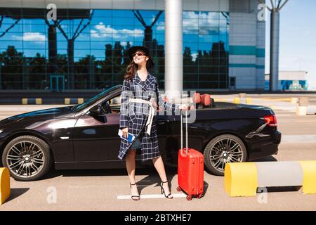 Jolie fille souriante en lunettes de soleil et chapeau noir tenant sur les talons passeport avec billet d'avion et valise rouge près de l'aéroport avec cabriolet Banque D'Images