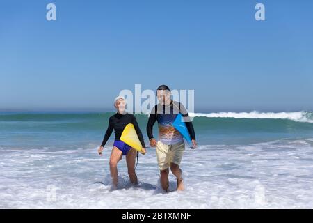 Couple caucasien senior appréciant le temps à la plage par une journée ensoleillée, marchant et tenant des planches de surf avec la mer en arrière-plan Banque D'Images