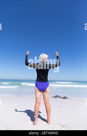 Femme caucasienne senior appréciant du temps à la plage par une journée ensoleillée, debout et de la flexion de ses biceps avec la mer en arrière-plan Banque D'Images