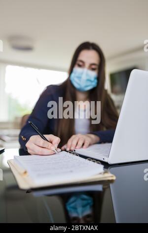 Portrait d'une jeune femme avec masque de protection chirurgical, assise sur le bureau devant son ordinateur portable et prenant des notes. Concept d'apprentissage en ligne. Banque D'Images