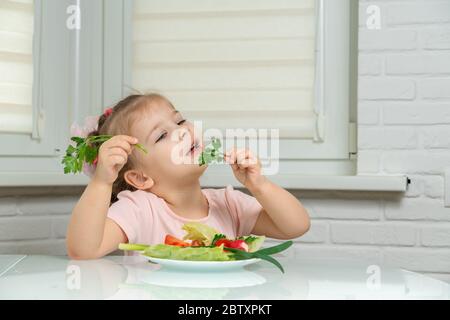 fille de 4 ans assise dans la cuisine à la table et manger des légumes, dans les mains d'une branche de persil. règles de nutrition préscolaire Banque D'Images