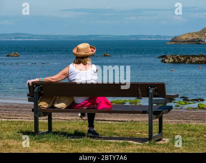 North Berwick, East Lothian, Écosse, Royaume-Uni, 28 mai 2020. Assouplissement des restrictions de verrouillage : le gouvernement écossais étant sur le point d'annoncer la levée de certaines restrictions de verrouillage aujourd'hui, des signes indiquent déjà que les choses commencent à revenir à la normale dans la populaire station balnéaire. Une femme portant un chapeau de paille se trouve sur un banc pour admirer la vue et se détendre par temps chaud Banque D'Images